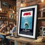 Framed Jaws poster on a brick wall in a cozy cafe with people at a table in the background and a popcorn cone on the counter nearby.