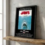 Framed Jaws movie poster leaning on a wooden shelf beside a concrete wall.