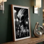Framed black-and-white poster of a gray alien holding a beer bottle, advertising UFO IPA on a wooden shelf.