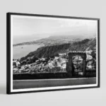 Framed black and white printable art of Taormina and the Bay of Naxos, Sicily—a stone gateway framing the coastal vista below, captured by Mark Marsh and displayed in a modern interior.