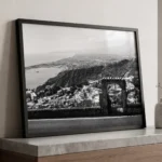 Taormina Bay black and white digital download print in a black frame, styled in a living room or study with natural textures, capturing the Sicilian coastal panorama by Mark Marsh.