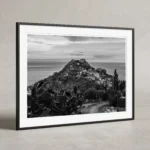 Framed black and white printable art of Castello di Taormina on Monte Tauro, Sicily—an elevated view over prickly pear cacti toward the Ionian Sea, captured by Mark Marsh and displayed in a modern interior.