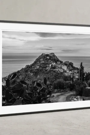 Framed black and white printable art of Castello di Taormina on Monte Tauro, Sicily—an elevated view over prickly pear cacti toward the Ionian Sea, captured by Mark Marsh and displayed in a modern interior.