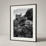Framed black and white printable art of Chiesa di San Nicolò in Savoca, Sicily—viewed across a steep valley with Mediterranean trees and tiled roofs below the crowning church, captured by Mark Marsh and displayed in a modern interior.
