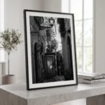 Framed black and white printable art of a narrow DIOR alleyway in Taormina, Sicily—glass display case framed by stone walls and iron lanterns, captured by Mark Marsh and displayed in a modern interior.