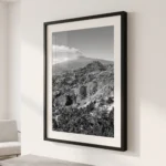 Framed black and white printable art of Mount Etna venting volcanic smoke, viewed from Taormina heights in Sicily—layered valleys and settlements beneath the peak, captured by Mark Marsh and displayed in a modern interior.