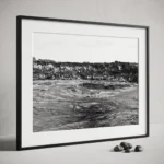 Framed black and white printable art of a basalt lava quarry on Mount Etna, Sicily—towering rock wall above a dusty excavated basin with tyre tracks, captured by Mark Marsh and displayed next to some decorative stones.