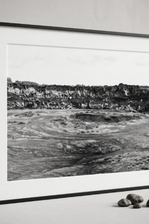 Framed black and white printable art of a basalt lava quarry on Mount Etna, Sicily—towering rock wall above a dusty excavated basin with tyre tracks, captured by Mark Marsh and displayed next to some decorative stones.