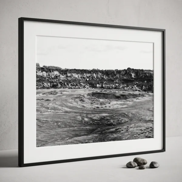 Etna Quarry black and white printable art printed at A2 size Framed black and white printable art of a basalt lava quarry on Mount Etna, Sicily—towering rock wall above a dusty excavated basin with tyre tracks, captured by Mark Marsh and displayed next to some decorative stones.