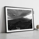 Framed black and white printable art of Mount Etna beneath a storm ceiling, viewed from a stone terrace above Taormina, Sicily—volcanic steam and roiling clouds, captured by Mark Marsh and displayed in a modern interior.