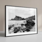 Isola Bella black and white digital download print in a black frame, styled with natural stone textures, capturing the Sicilian coast by Mark Marsh.