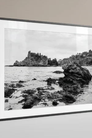 Isola Bella black and white digital download print in a black frame, styled with natural stone textures, capturing the Sicilian coast by Mark Marsh.