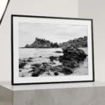 Framed black and white printable art of Isola Bella from the shoreline below Taormina, Sicily—volcanic rocks in the foreground and the islet across a calm bay, captured by Mark Marsh and displayed in a modern interior.