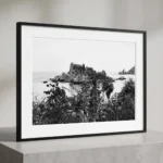 Isola Bella through the wild black and white digital download print in a black frame, styled in a room with natural stone textures, capturing the Sicilian coast by Mark Marsh.