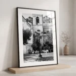 Framed black and white printable art of Piazza IX Aprile in Taormina, Sicily—an ornate lamppost on checkerboard tiles with Chiesa di San Giuseppe and Monte Tauro above, captured by Mark Marsh and displayed in a modern interior.