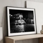 Framed black and white printable art of Piazza Sant'Antonino in Castelmola, Sicily at night—mosaic tiles, glowing café facade, and rustic stone buildings, captured by Mark Marsh and displayed in a modern interior.