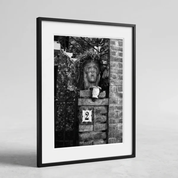 Plant pot head at number 2 art print by Mark Marsh in small wooden frame Framed Taormina Testa di Moro black and white digital wall art print by Mark Marsh, showing the sculpted ceramic face, trailing plant leaves, and rose-bordered house number tile detail.