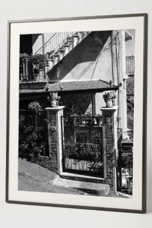 Framed black and white printable art of twin Sicilian Testa di Moro planters guarding a residential entrance in Castelmola—stone pillars, iron gate, and staircase shadow, captured by Mark Marsh.