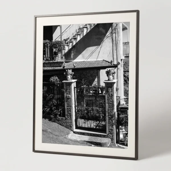 Plant pot heads on Sicilian house digital download by Mark Marsh in brown frame Framed black and white printable art of twin Sicilian Testa di Moro planters guarding a residential entrance in Castelmola—stone pillars, iron gate, and staircase shadow, captured by Mark Marsh.